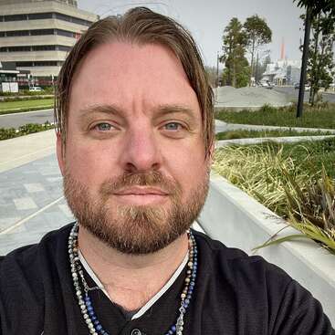 A man with light brown hair and a beard takes a selfie outdoors, wearing beaded necklaces. Buildings, greenery, and a pathway are visible in the background.