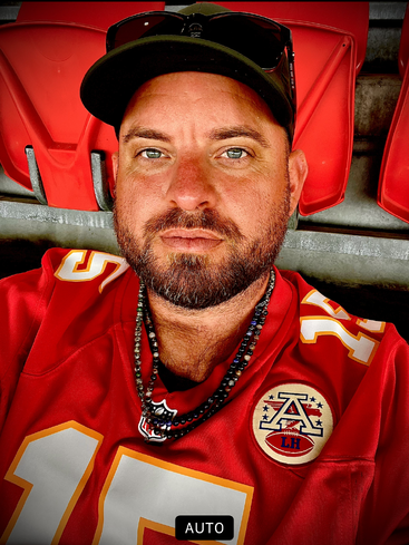 A man with a beard, wearing a red football jersey, black cap, and sunglasses, sits in red stadium seats, looking directly into the camera for a selfie.