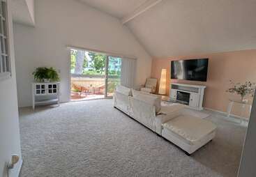This spacious living room features a vaulted ceiling, neutral colors, cozy white furniture, a modern fireplace, large TV, patio access, and abundant natural light.