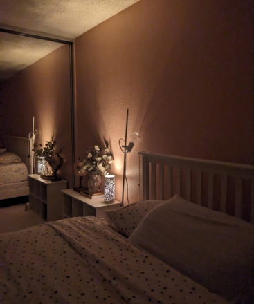 Um quarto aconchegante com iluminação suave, flores decorativas na mesinha de cabeceira, roupa de cama estampada e um grande espelho que reflete a atmosfera acolhedora, convidativa e tranquila.