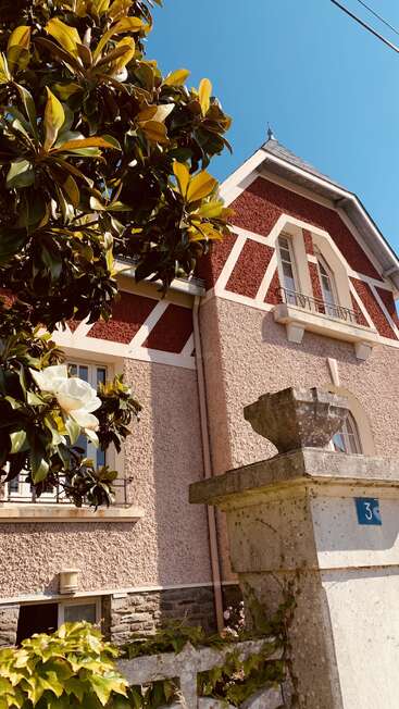 A charming house with textured pink walls, red accents, and white trim, framed by a magnolia tree and blue sky. Stone pillar with number 3.