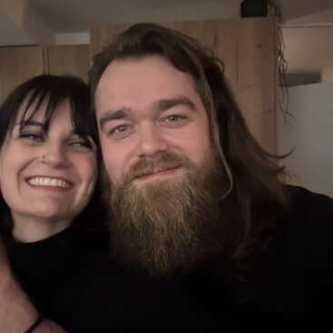 Eine lächelnde Frau mit dunklen Haaren und ein Mann mit langen Haaren und Vollbart posieren eng beieinander in einem Haus und wirken glücklich und genießen die Gesellschaft des anderen.