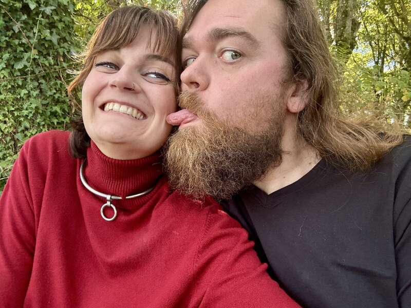 Uma mulher sorridente em um suéter vermelho posa para uma selfie com um homem barbudo que brinca com a língua na bochecha dela, ambos parecendo felizes ao ar livre.