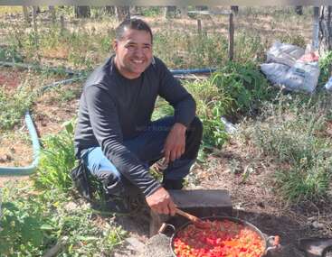 A man squats outdoors, smiling while cooking a colorful dish in a pan over an open fire, surrounded by greenery, plants, and gardening supplies.
