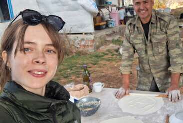 A woman takes a selfie with a smiling man who is rolling dough outside on a table. Various kitchen supplies and food items are spread around them.