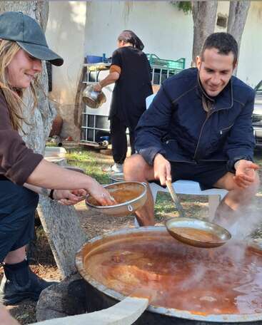 Two people are sitting outdoors, smiling as they prepare food together over a large steaming pot. Another person works in the background, enhancing the communal atmosphere.