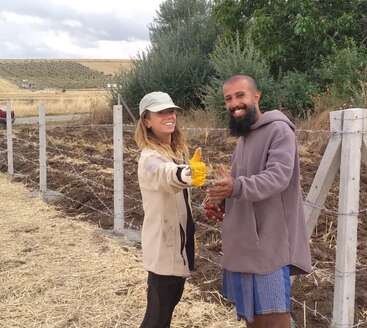 Two people are standing by a fence in a rural area, smiling and appearing happy. One gives a thumbs up, both wear casual outdoor clothing.