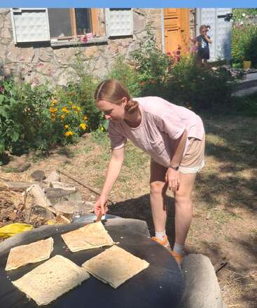 A woman cooks flatbreads on a large outdoor pan in a garden. Flowers bloom behind her, and a young child stands near the house watching.