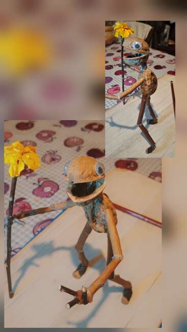 This image shows a quirky, handmade creature sculpture made from nuts, sticks, and googly eyes. It holds a yellow flower and stands on a patterned table.