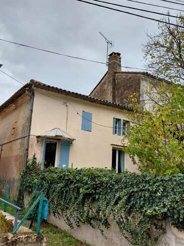 A rustic house with cream walls and blue shutters, surrounded by lush ivy-covered fencing, stands under a cloudy sky, with trees and power lines overhead.
