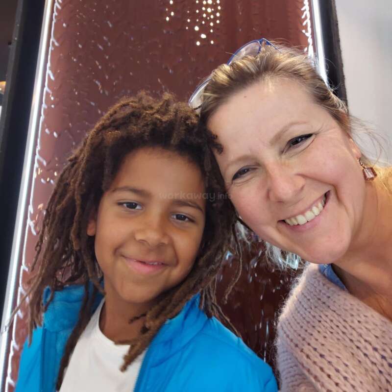 A smiling woman and a young boy with dreadlocks pose closely together for a cheerful selfie. The background features a dark, patterned wall with light accents.