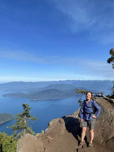 A woman stands smiling at a scenic mountain viewpoint, overlooking deep blue water and islands, with a clear sky above. She’s dressed in hiking gear.