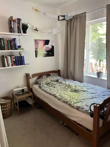 A cozy bedroom with a wooden bed, gray patterned bedding, bookshelves, wall art, string decorations, a large window with curtains, a nightstand, and a laundry basket.