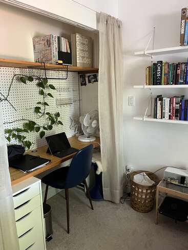 This cozy home office features a wooden desk, blue chair, pegboard with plants, bookshelves, fan, wicker basket, and curtains, creating a tidy and inviting workspace.