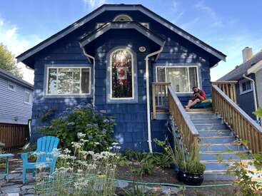 A charming blue house with white trim, wooden stairs, a garden with flowers, a Canadian flag in the window, and a person sitting on the porch.