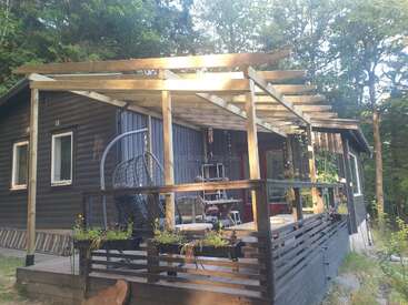 Une cabane en bois confortable est dotée d'une pergola partiellement construite au-dessus de sa terrasse, d'une chaise suspendue, de plantes en pot et de sièges extérieurs, le tout entouré d'une forêt verdoyante.