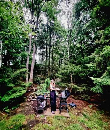 Une femme se tient près d'un barbecue dans une forêt verdoyante. Elle sourit, entourée de grands arbres, de la nature et d'un cadre extérieur rustique.