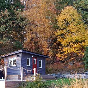 Une petite cabane bleue avec une porte rouge se trouve devant des arbres d'automne éclatants, entourée de nature, de rochers et de verdure lors d'une journée paisible.