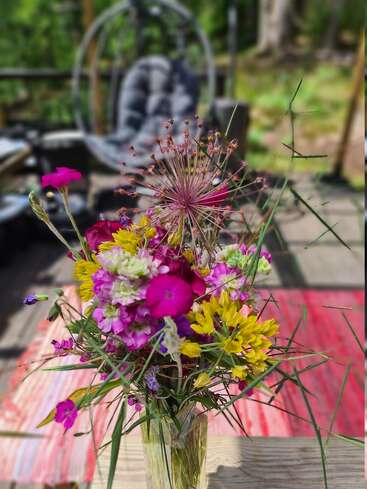 Un bouquet vibrant de fleurs sauvages dans un vase en verre est posé sur une table en bois à l'extérieur, avec un arrière-plan flou représentant un fauteuil suspendu et rembourré.