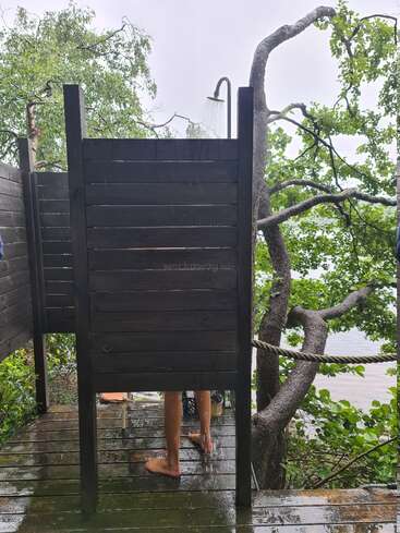 Une personne se douche en plein air dans une cabine en bois, entourée d'arbres verdoyants, avec des gouttes d'eau visibles. Le cadre semble naturel, paisible et rafraîchissant.