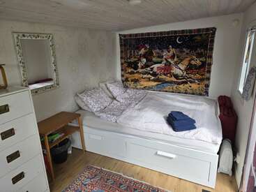 Une chambre confortable avec un lit blanc, une literie florale, une tapisserie colorée représentant des cavaliers, une petite table en bois, une commode, un tapis, un miroir et de la lumière naturelle.
