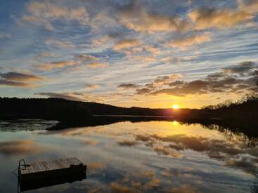 Un lac tranquille reflète un magnifique coucher de soleil, avec des rayons de soleil dorés traversant les nuages. Un quai en bois flotte dans l'eau calme, entouré d'arbres et de collines sereines.