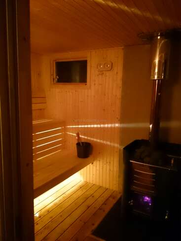 Cette image montre un sauna en bois confortable avec un éclairage chaleureux, un banc, un seau, une petite fenêtre et un poêle traditionnel chauffant des pierres dans le coin.