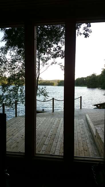 À travers de grandes baies vitrées, une terrasse en bois surplombe un lac tranquille entouré d'arbres, la lumière douce du soleil illuminant le cadre paisible et naturel de l'extérieur.