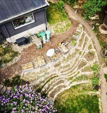 Cette image aérienne montre un jardin en terrasse avec des plates-bandes surélevées, des chemins en pierre, une maison sombre, des poubelles vertes, un banc extérieur, des fleurs et la verdure environnante par une journée ensoleillée.