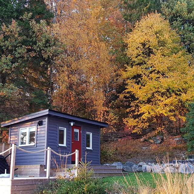 Une petite cabane bleue avec une porte rouge se trouve devant des arbres d'automne éclatants, entourée de nature, de rochers et de verdure lors d'une journée paisible.