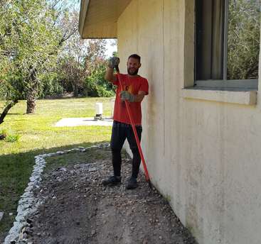 A man stands outside a house, holding a long pole, wearing gloves and a red t-shirt, with a garden hose and trees visible in the background.