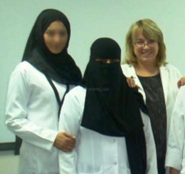 The image shows three women in white lab coats standing together, with two wearing headscarves and one wearing a niqab, posing for a photo.