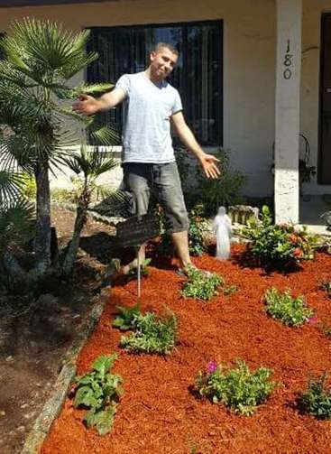 A man stands in a garden with red mulch, gesturing to a palm tree, wearing a grey t-shirt and shorts, in front of a house with a white pillar.