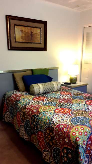 The image depicts a cozy bedroom with a vibrant bedspread, a framed picture of a dragonfly, and a bedside table with a lamp, exuding a warm and inviting atmosphere.