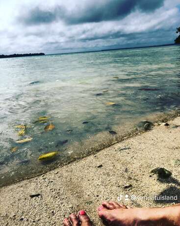Das Bild zeigt eine ruhige Strandszene mit den Füßen eines Menschen im Sand, der unter einem wolkenverhangenen Himmel auf ein Gewässer blickt, und ein Wasserzeichen, das anzeigt, dass es von der Insel Vanuatu stammt.