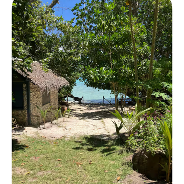 A thatched-roof hut sits among lush green trees near a sandy path leading to clear blue water, bathed in bright sunlight, creating a tropical paradise atmosphere.