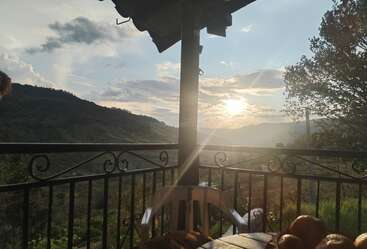 Una hermosa vista desde el balcón al atardecer, con vistas a verdes colinas y un paisaje sereno. Una mesa con fruta y una silla invitan a relajarse y disfrutar tranquilamente.