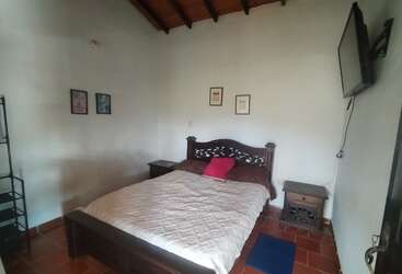 Esta acogedora habitación cuenta con una cama de madera con una colcha blanca y almohadas rosas, mesitas de noche, cuadros decorativos, estanterías, suelo de baldosas y un televisor montado en la pared.