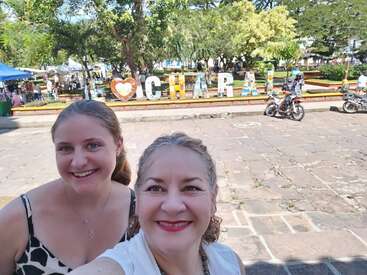 Duas mulheres sorridentes tiram uma selfie em um parque ensolarado. Atrás delas, aparecem letras coloridas que formam a palavra “CHARAL” e um coração. Dá pra ver pessoas e motos.