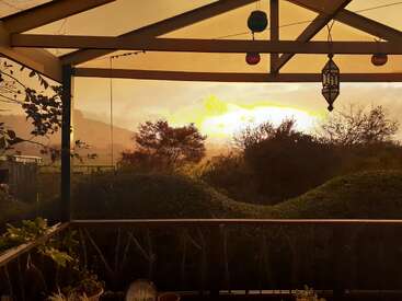 A warm sunset view from a covered patio, overlooking lush bushes and silhouetted trees, with hanging decorations creating a cozy and tranquil outdoor scene.