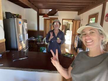 Two people smiling in a cozy kitchen, both making peace signs. Sunlight enters from the door. The atmosphere is cheerful, relaxed, and casual with simple home decor.