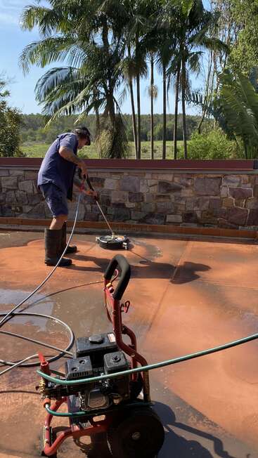 A man in rubber boots uses a pressure washer to clean a patio, surrounded by lush greenery and palm trees under a bright, sunny sky.