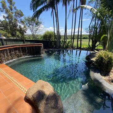 Uma bela piscina tropical com água azul clara, uma cachoeira de pedra, palmeiras, vegetação exuberante, piso de ladrilho vermelho e uma grande rocha sob o sol forte.