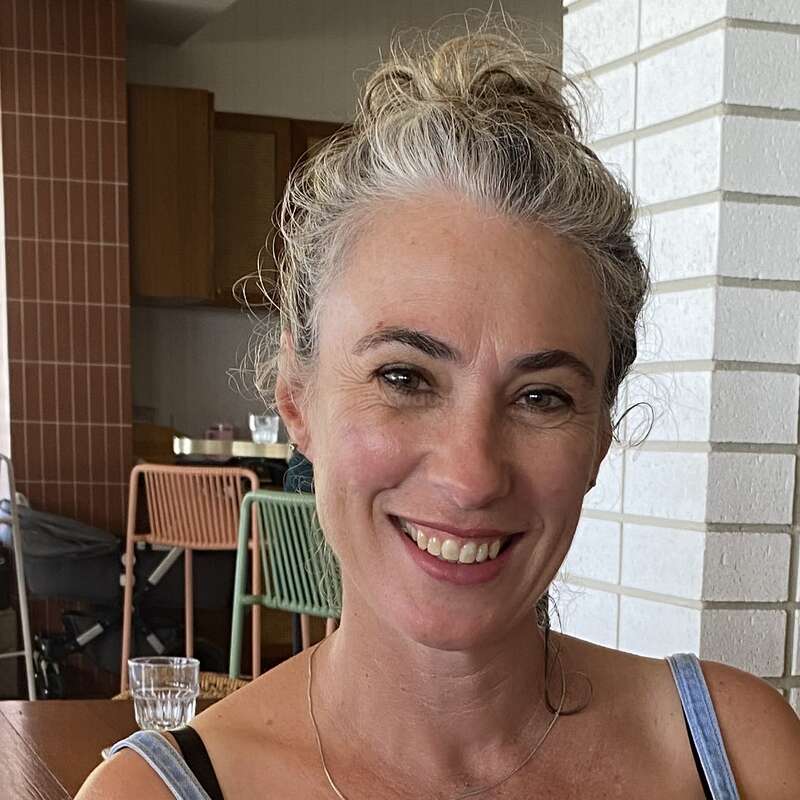 A woman with curly, gray hair in a bun is smiling indoors. She wears a blue tank top and necklace, sitting near white brick and wooden furniture.