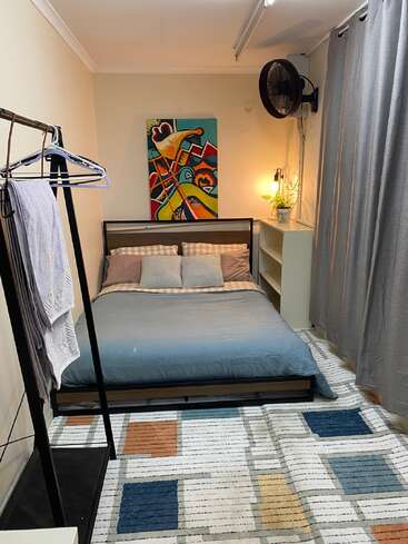 Un acogedor dormitorio con cama de matrimonio, arte abstracto en la pared, una alfombra de colores, estanterías, una maceta, ropa colgada y cortinas grises. Iluminación cálida y acogedora.