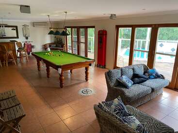 Amplia sala de juegos con mesa de billar, sofás de ratán, zona de bar y grandes ventanales con vistas a la piscina. La cálida iluminación y el suelo de baldosas crean un ambiente acogedor.