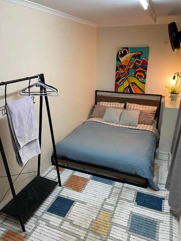 Un acogedor dormitorio con cama de matrimonio, ropa de cama azul, alfombra con motivos geométricos, perchero, colorido arte abstracto, iluminación tenue y una pequeña planta en la estantería.