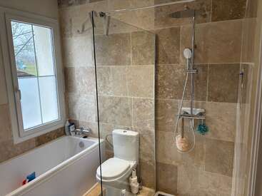 L'image représente une salle de bains avec une baignoire, des toilettes et une douche, avec des carreaux beiges sur les murs et une fenêtre avec un cadre blanc.