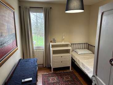 L'image représente une petite chambre confortable avec un lit simple, une commode blanche et une fenêtre, agrémentée d'un tapis, d'une lampe et d'œuvres d'art sur les murs, créant une atmosphère sereine.