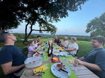 L'image représente une famille de huit personnes prenant un repas à une longue table en plein air, entourée d'arbres et d'un paysage serein.